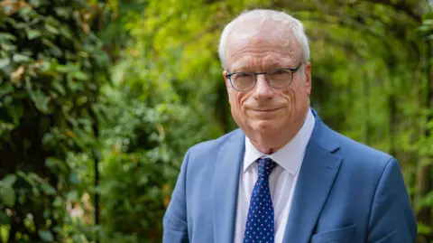 PA Lord Chris Smith lleva traje azul y corbata y se encuentra en jardines frondosos.