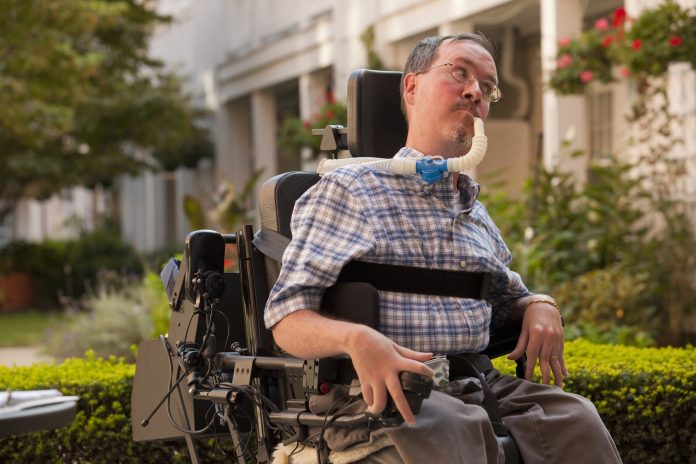 Hombre con distrofia muscular de Duchenne sentado en una silla de ruedas motorizada usando un controlador de potencia con manos degeneradas. Dyne Therapeutics está desarrollando un tratamiento para algunas personas con DMD.
