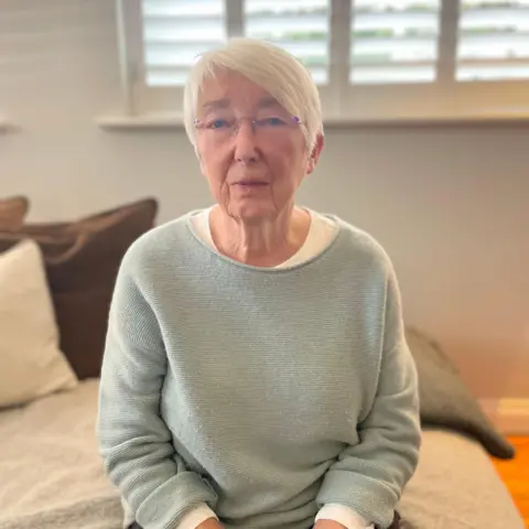 Pauline Collier sentada en una cama. Tiene el pelo corto y blanco y lleva un suéter azul pálido.