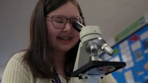 Una chica de largo cabello castaño y gafas baja las escaleras de un microscopio en una clase de biología.