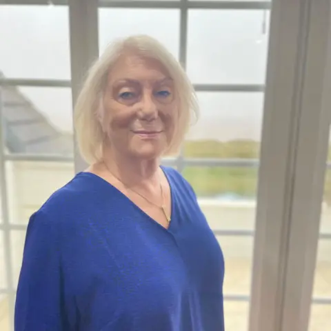 Carolyn lleva una camisa azul y está parada junto a la ventana de su casa mirando a la cámara.