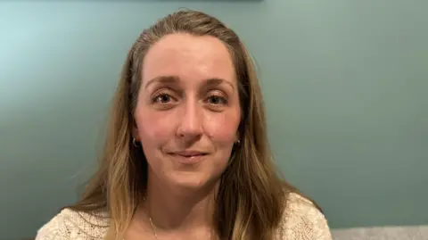 Molly Smith / BBC Celestina Mahovic tiene el pelo largo, castaño claro y sonríe. Lleva un suéter blanco y está sentada frente a una pared verde azul.