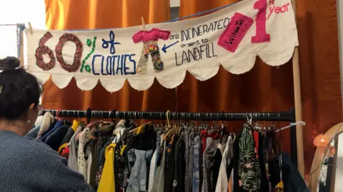 Molly Smith / BBC Había un perchero con muchos tipos diferentes de ropa colgada en él. Tiene un estampado con palabras bordadas que dicen: El 60% de toda la ropa se incinera o se envía al vertedero durante el primer año. Hay una mujer arrastrando ropa y se la puede ver desde atrás.