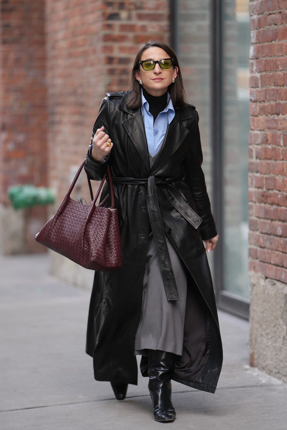 nueva york, 12 de febrero de nueva york, un invitado lleva un corte de pelo, gafas de sol de color verde, pendientes de plata, una bolsa tote de cuero tejida de color rojo borgoña bottega venita, una gabardina larga con cinturón de cuero negro sobre una camisa azul con botones y rectas, botas negras, zapatos de cuero durante la semana de la moda de New York Zanghkov 12 de febrero de 2026 en la ciudad de Nueva York, fotografía de edward berthelotgetty images