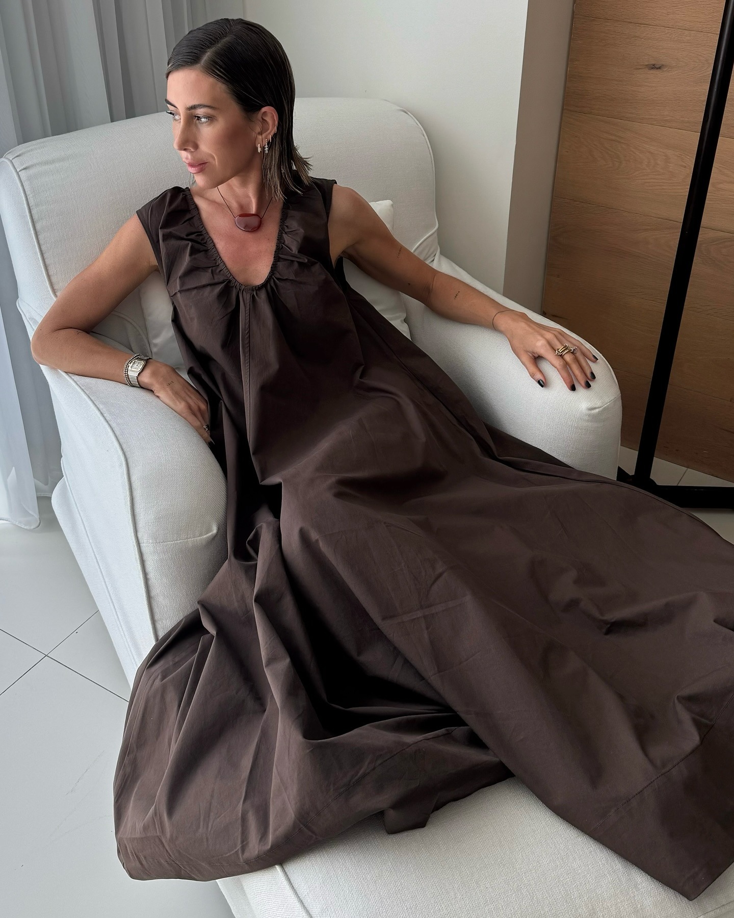 La influencer británica Hannah Lewis pone en una silla de lino blanco con un traje maxi de cuello abierto con frun marrón y un collar de cordón negro con una piedra roja.