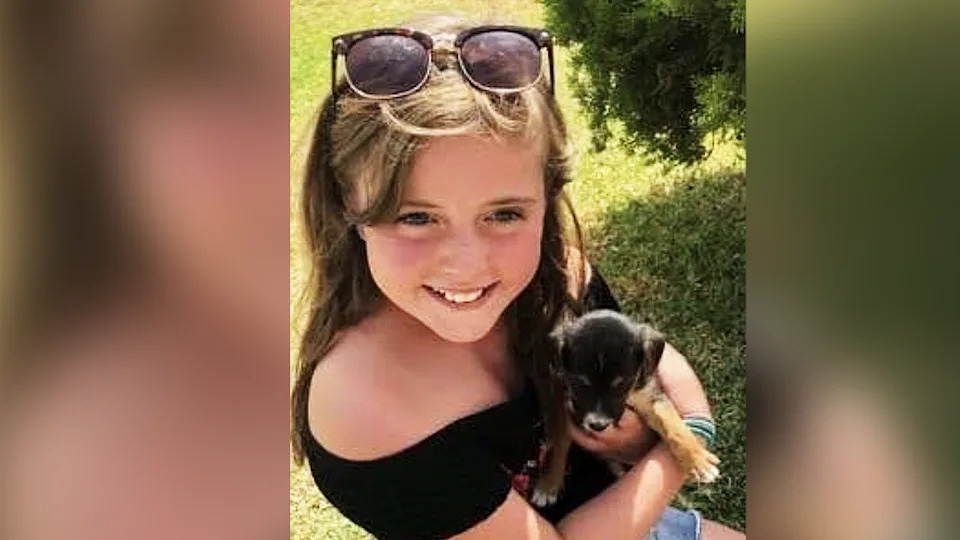 Nia, de unos nueve años. Lleva una parte superior y pantalón corto negro y lleva gafas de sol en la cabeza. Tiene el pelo largo y castaño claro. Lleva un cachorro negro y marrón. Ella sonríe directamente, pero la foto está hecha desde arriba, así que no cabe en la cámara.