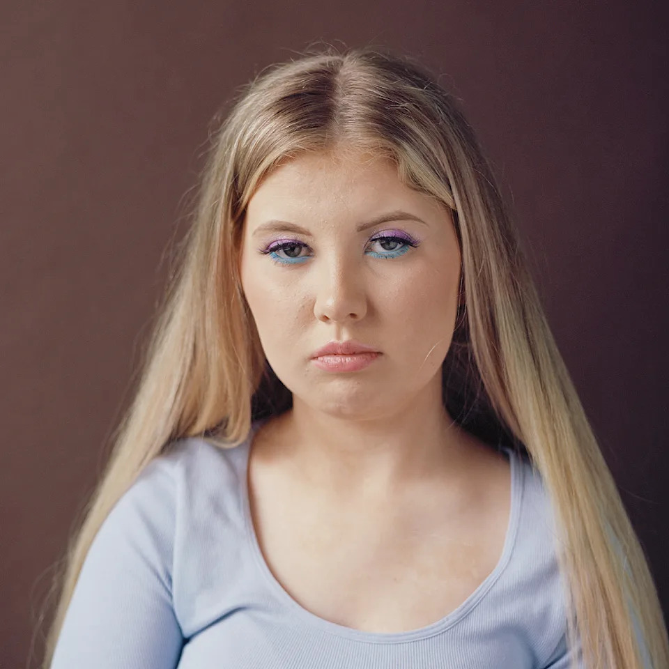 Imagen de la cabeza y los hombros de una chica con el pelo largo y rubio. Lleva una parte superior de algodón azul claro y tiene un maquillaje de ojos sorprendente, con un delineador de ojos azul eléctrico debajo y sombra de ojos de color violeta claro en los párpados. Ella mira a la cámara sin sonreír.