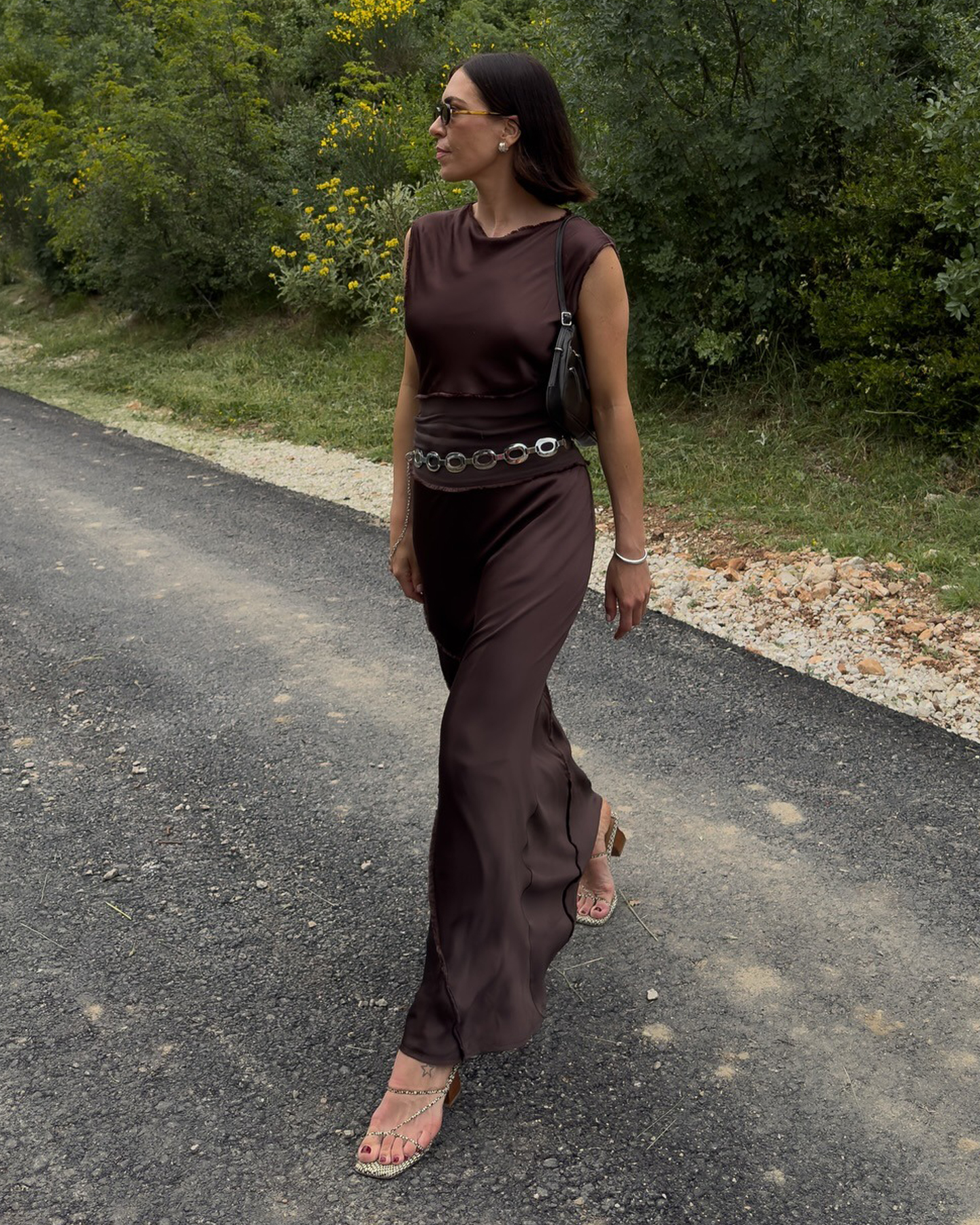 La influencer de estilo británico Jessica Skye pone con un traje maxi de seda de cuello marrón con un cinturón de cadena plateada y sandalias de piel de serpiente de tirantes.