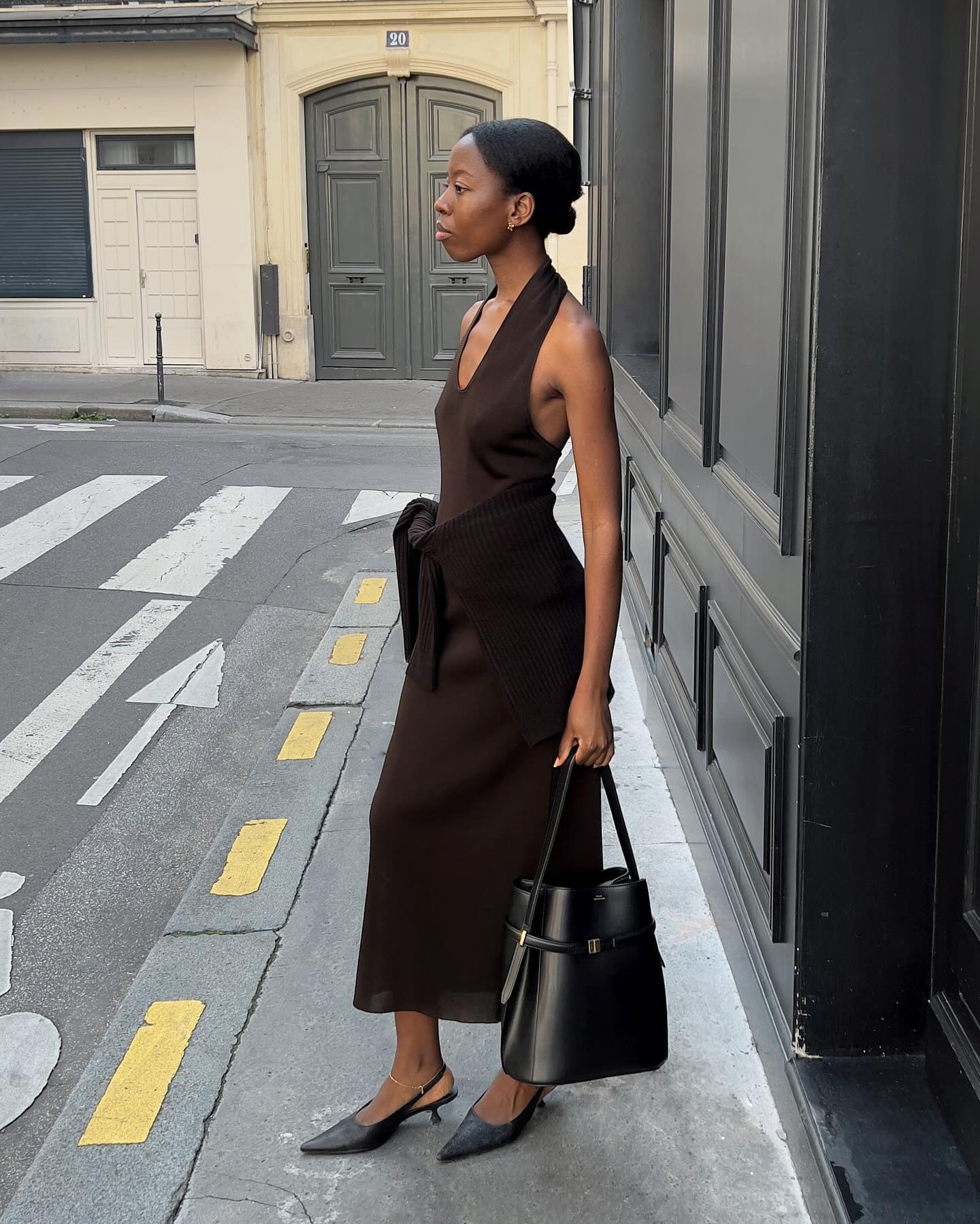 La influencia de estilo europeo Sylvie Mus pone en una acera de París con un traje marrón con cuello halter con un jersey atado a la cintura, una bolsa Toteme y unos tacones negros.