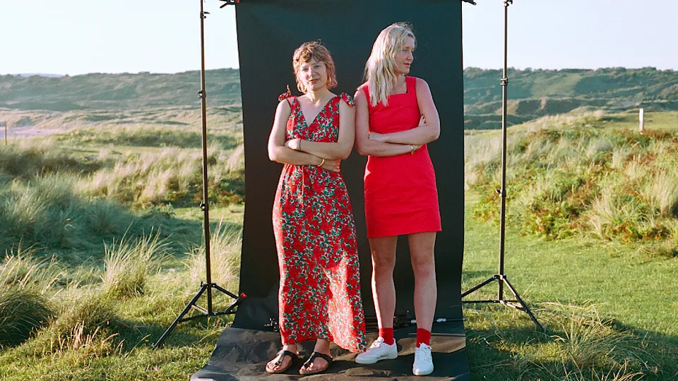 Clémentine Schneidermann y Charlotte James están una junto al otro frente a un fondo de fotografía negro que se encuentra en un paisaje salvaje de túmulos herbosos y pequeñas colinas al fondo. Clémentine se encuentra a la izquierda de la imagen con un vestido largo de color naranja sin mangas con un dibujo gris y blanco, sandalias de tanga de cuero y una pulsera. Tiene el pelo rojo teñido hasta el cuello y sonríe a la cámara. Charlotte lleva un traje ajustado a medio muslo de color naranja brillante con zapatillas blancas y calcetines naranjas. Tiene el pelo largo teñido de rubio peróxido y mira hacia la derecha de la imagen.