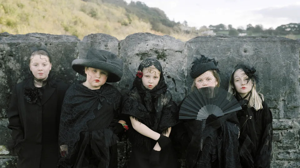 Cinco chicas blancas, de entre ocho y diez años aproximadamente, van vestidas con elaborados trajes negros, velos y sombreros. Uno tiene un ventilador. La ropa está llena de encaje y plumas y tiene un aspecto victoriano en general. Se encuentran frente a una pared que es más alta que ellos. Detrás hay una colina hierba desenfocada y casas a lo lejos.